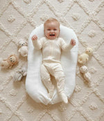 Baby lying on a white cushion with soft toys around on a textured beige surface