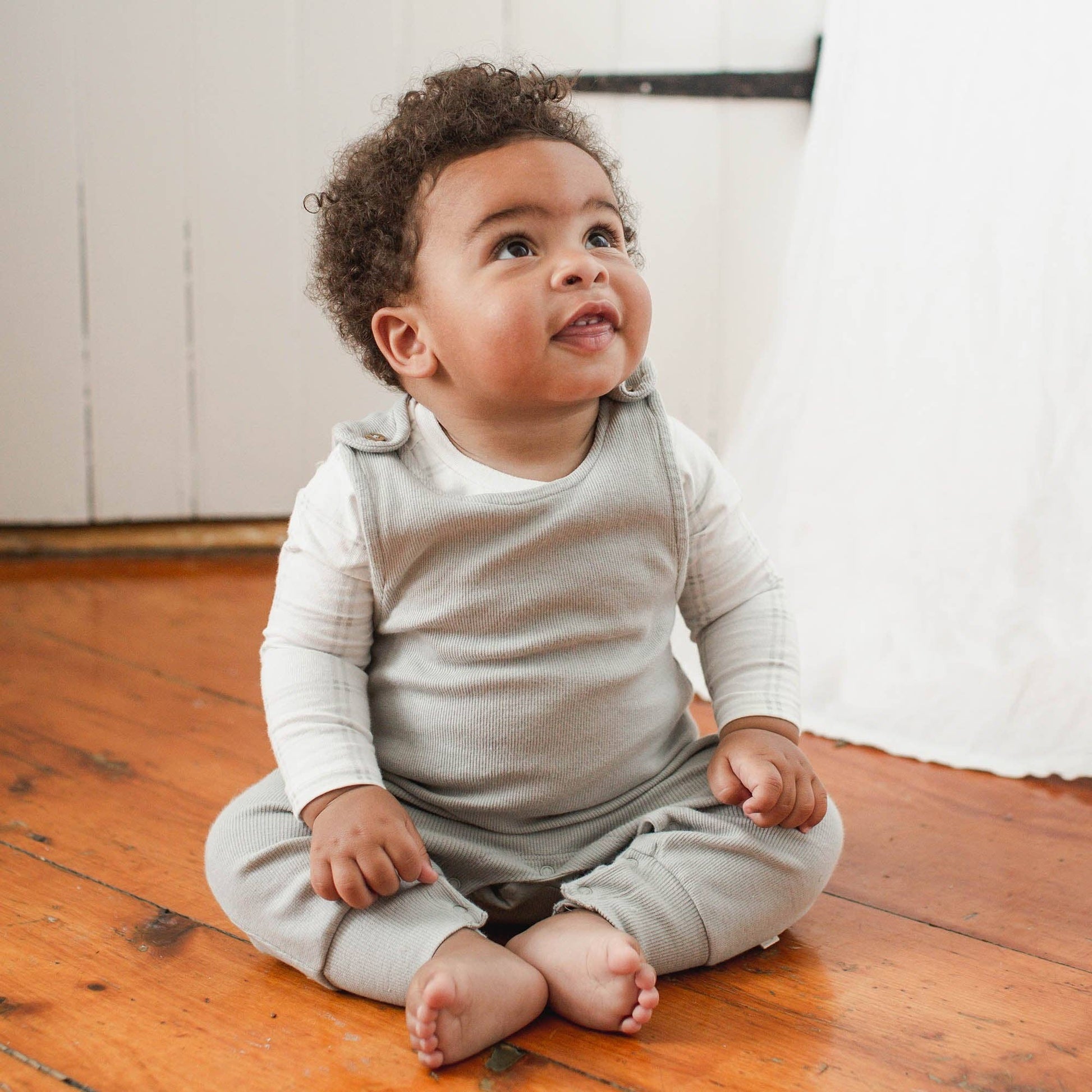 Baby sitting on a wooden floor wearing a light gray overalls with a white onesie.