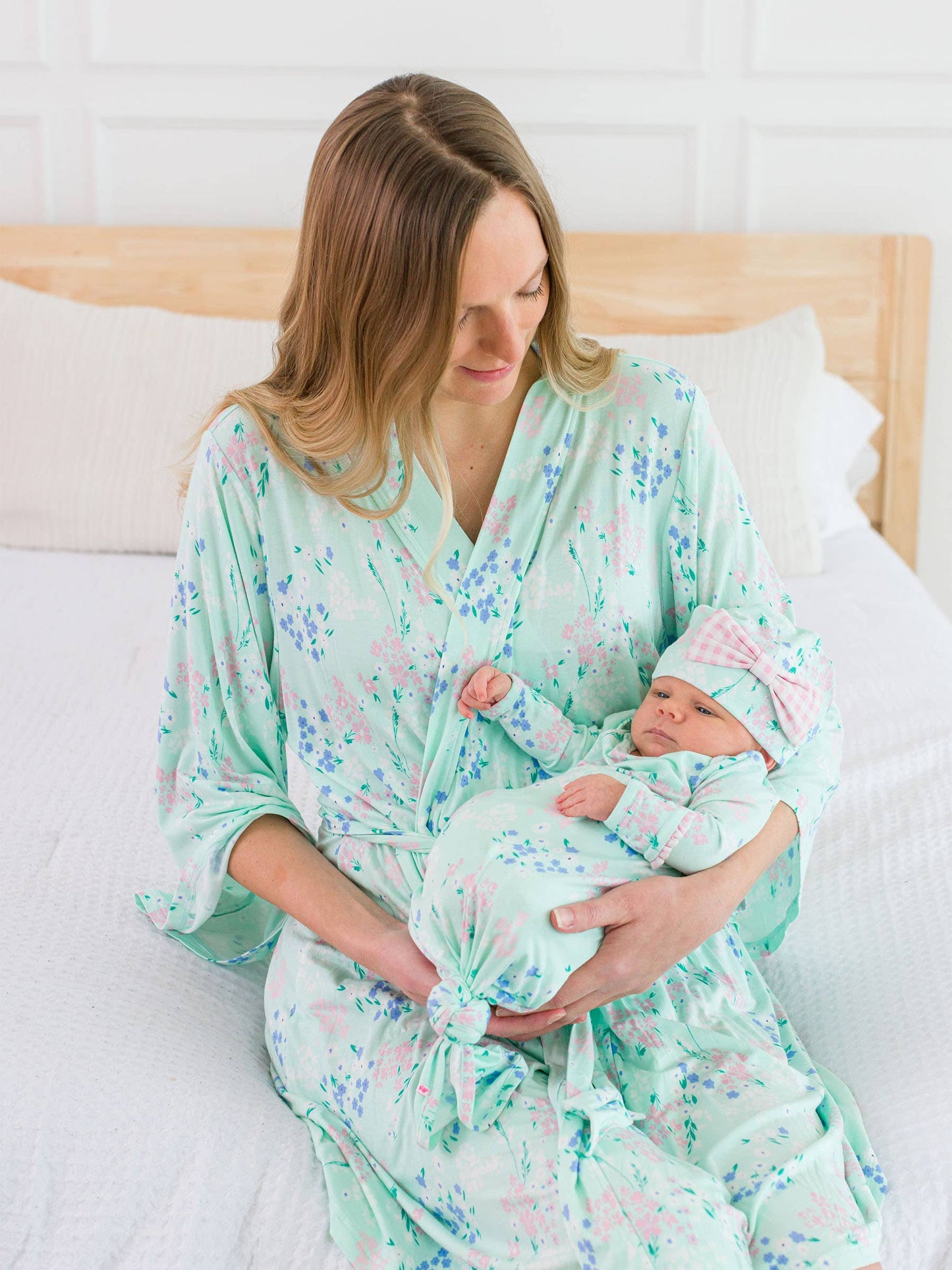 Woman in a floral robe holding a baby wrapped in a matching knotted gown on a bed.