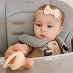 Baby in a high chair wearing a bow headband and holding a pacifier self feeder