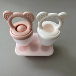 Pink and white baby self feeder pacifiers with a stand on a gray background
