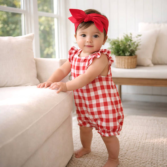 Baby in a red checkered outfit with a large bow standing on a light-colored couch.