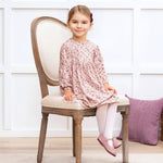 Young girl in a floral dress sitting on a wooden chair with a white wall and purple pillow in the background.