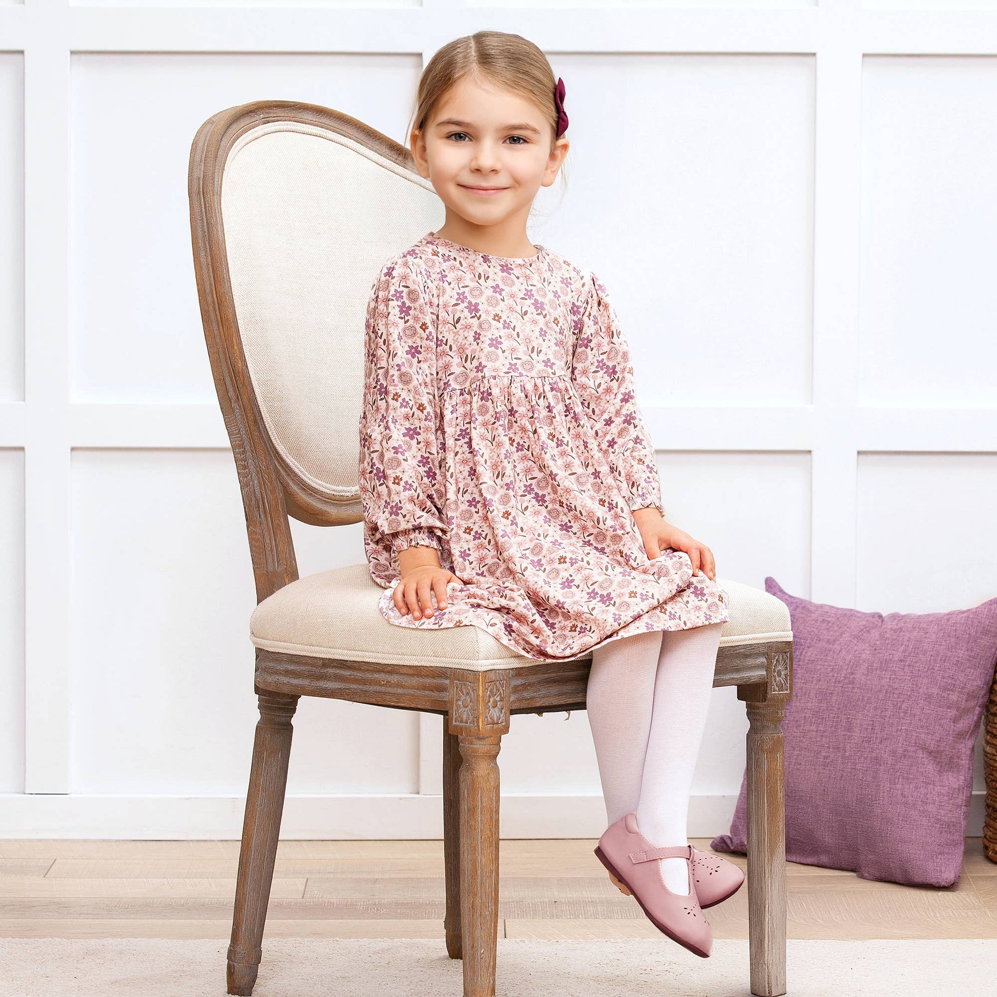 Young girl in a floral dress sitting on a wooden chair with a white wall and purple pillow in the background.