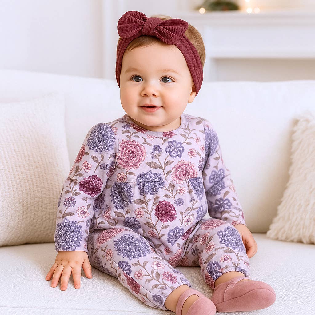 Baby wearing a floral onesie and headband sitting on a white couch.